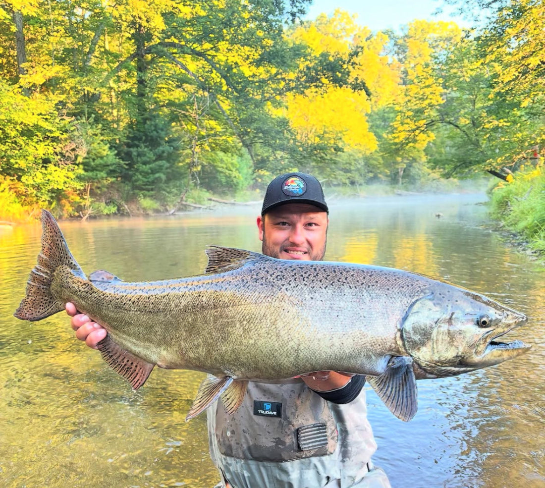 Man holding a large fish by a river with trees in the background